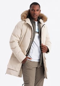 Béžová prošívaná parka s kapucí lemovanou kožešinou, s plastovým zipem a knoflíkovým zapínáním, a velkými předními kapsami. Model má na sobě olivově zelenou košili a bílé tričko.