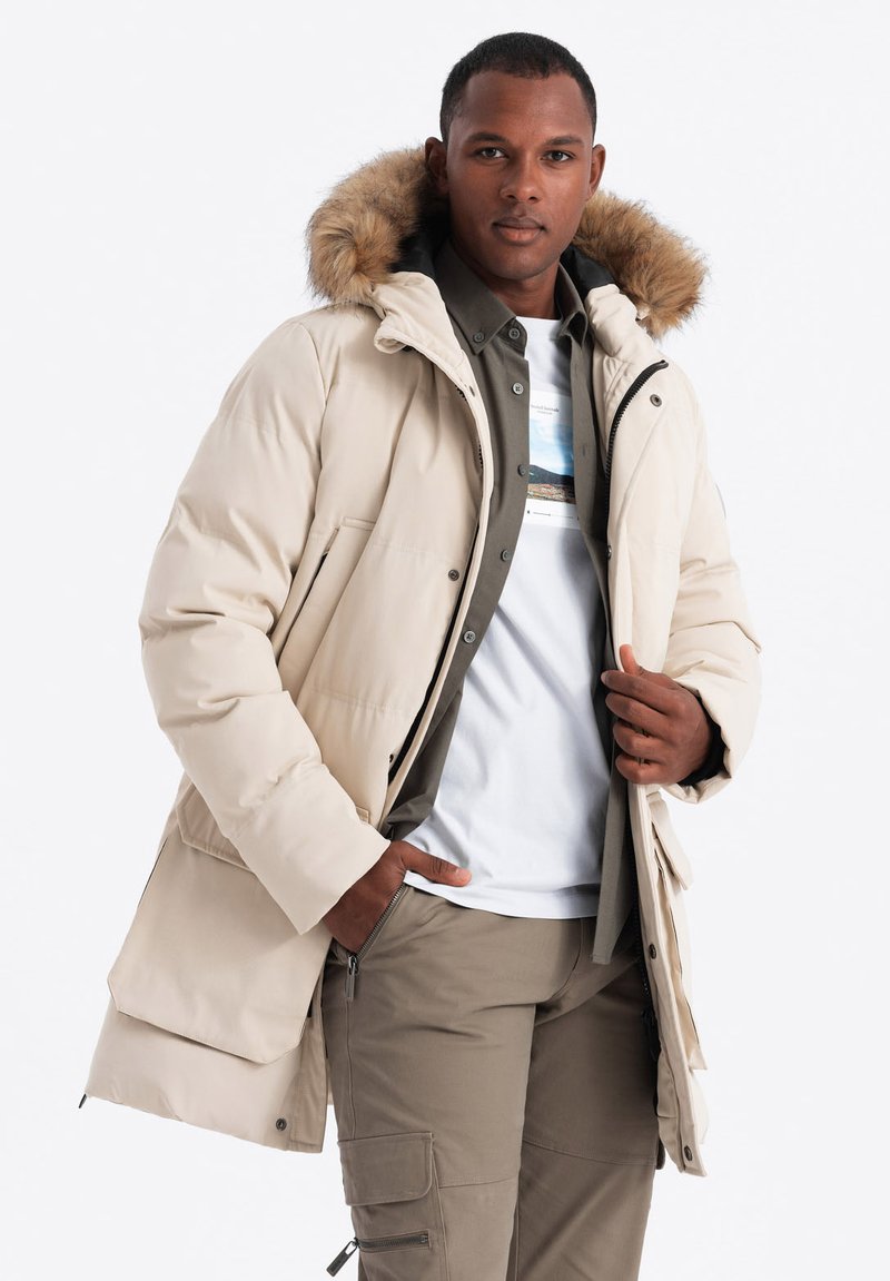 Béžová prošívaná parka s kapucí lemovanou kožešinou, s plastovým zipem a knoflíkovým zapínáním, a velkými předními kapsami. Model má na sobě olivově zelenou košili a bílé tričko.