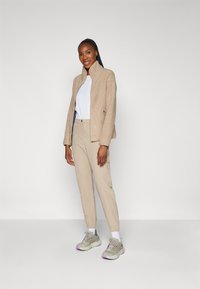 Giacca in pile beige, camicia bianca, pantaloni beige e sneakers grigio chiaro con accenti lavanda. Il modello è in piedi con un'espressione neutra.
