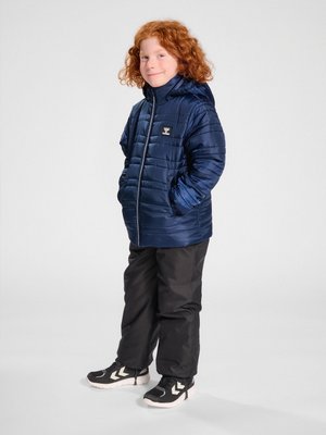 Dziecko z kręconymi rudymi włosami, ubrane w granatową puchową kurtkę, czarne spodnie i czarne trampki z białymi podeszwami, stojące na białym tle.