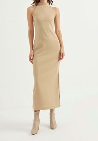 Woman wearing a sleeveless beige ribbed maxi dress with a high neckline and side slit, paired with beige ankle boots.