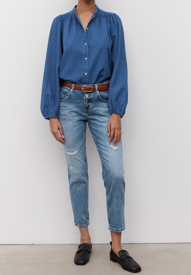 Femme portant une chemise bleue à manches longues boutonnée, un jean bleu usé avec une ceinture marron et des mocassins noirs, debout contre un mur blanc.