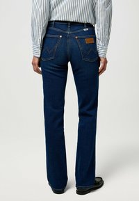 Jeans in denim blu con taglio dritto, dotati di due tasche posteriori con un motivo a zigzag e una patta con logo in pelle. Indossati con una camicia a righe.