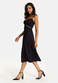 Black sleeveless dress with a fitted bodice and side cutout. Flowy skirt extends to mid-calf. Paired with black high-heeled shoes.