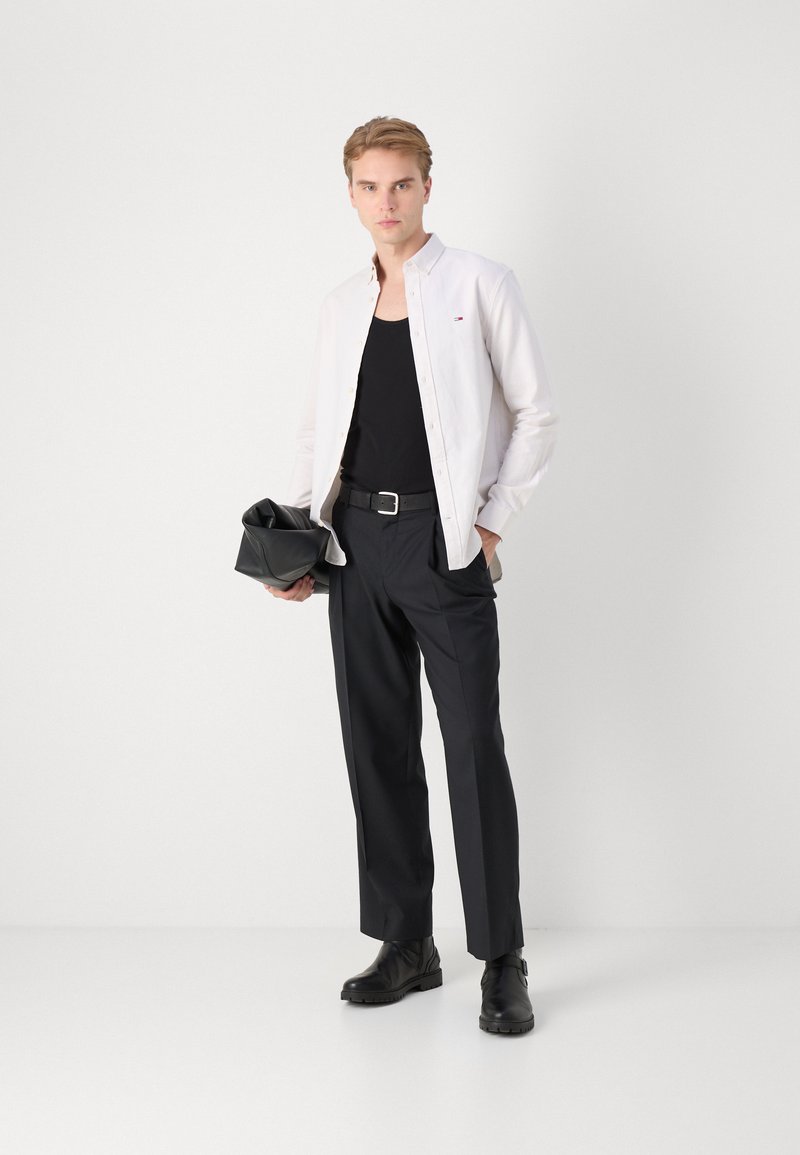 Camisa blanca de manga larga, top negro sin mangas, pantalones negros a medida, botas de cuero negras, sosteniendo un bolso negro. Diseño simple, texturas suaves.