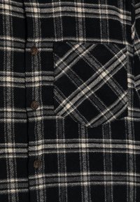 Camisa de franela a cuadros en blanco y negro con un bolsillo en el pecho, botones marrones y una textura suave con un patrón a cuadros.