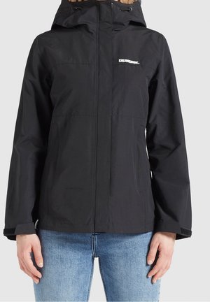 Zwarte capuchonjas met ritssluiting aan de voorkant en wit logo op de borst, gedragen over lichtblauwe spijkerbroek.