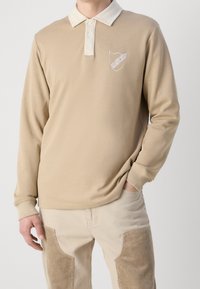 Beige långärmad polo med en kontrasterande vit krage, prydd med en sköldlogotyp broderad på vänster bröst. Mjuk textur.