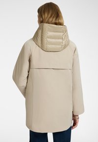 Donna che indossa un cappotto imbottito beige con cappuccio, con la schiena liscia e pantaloni scuri, in piedi di spalle contro uno sfondo bianco.