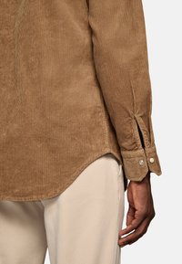 Bruine corduroy shirt met een gestructureerde afwerking, voorzien van een licht gebogen zoom en knoopmanchetten. De arm is naast lichtgekleurde broek gepositioneerd.