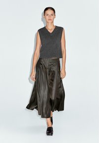 Grijze mouwloze gebreide top met een V-hals, gecombineerd met een lange, geplooide olijfgroene satijnen rok. Zwarte schoenen maken de outfit compleet.