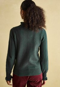 Grüner Pullover mit einem gerippten Kragen und Ärmeln, aus einem weichen Material. Besitzt eine strukturierte Oberfläche und eine körpernahe Passform. Kombiniert mit burgunderfarbenen Hosen.