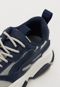 Tenis navy cu panouri din plasă, talpă din cauciuc, șireturi gri și branding. Dispone de un guler căptușit și o buclă la călcâi pentru a fi ușor de încălțat.