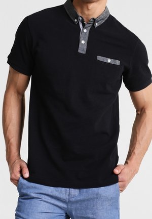 Man wearing black short-sleeve polo shirt with gray buttoned collar and chest pocket paired with light blue pants, hands in pockets.