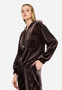 Sudadera de terciopelo marrón con cremallera frontal, dos bolsillos en el pecho, puños acanalados y capucha con cordón. Textura suave con un acabado brillante.