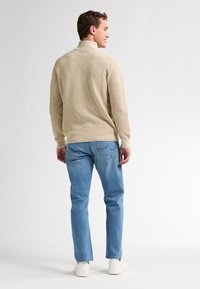 Beige ribbad tröja med hög krage, matchad med ljusblå jeans. Modellen bär vita sneakers, sedd bakifrån.