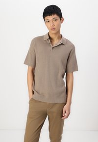 Polo à manches courtes, couleur marron clair, en tissu tricoté texturé, avec col et patte de boutonnage à trois boutons, associé à un pantalon beige.