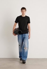 Zwart T-shirt met korte mouwen, lichtblauwe versleten jeans met grote scheuren, een zwarte tas dragen, staand op een neutrale achtergrond.