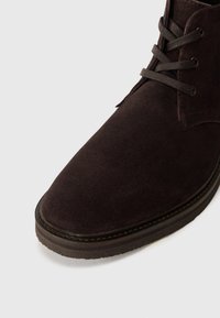 Gros plan d'une botte noire en daim marron foncé avec semelle cousue et œillets métalliques sur fond clair.