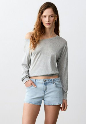 Jeune femme aux longs cheveux portant un pull gris clair à épaules dénudées et un short en jean clair, debout avec une main dans la poche.