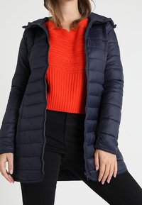 Marinblå pufferjacka med huva, med quiltat mönster, dragkedja framtill och lång skärning, bärs över en ljusröd stickad tröja.