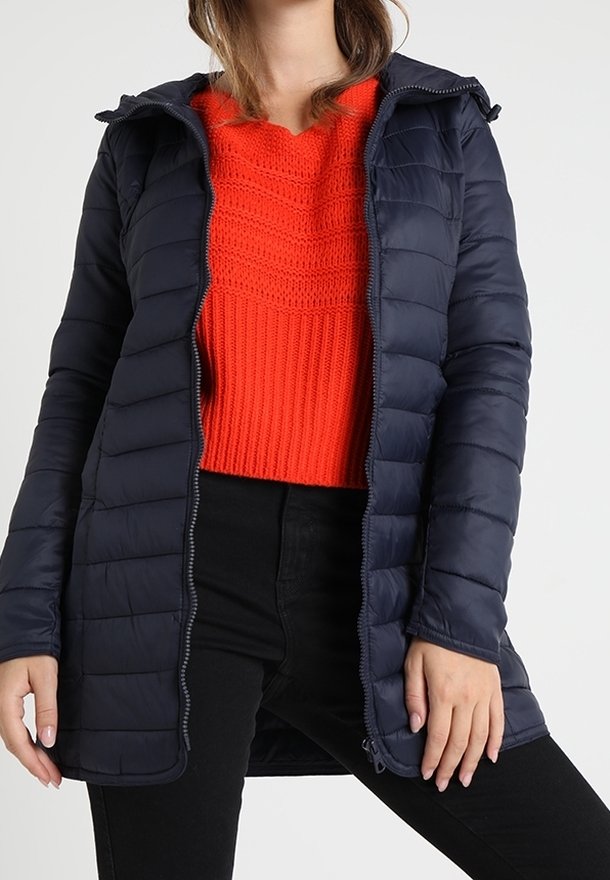 Marinblå pufferjacka med huva, med quiltat mönster, dragkedja framtill och lång skärning, bärs över en ljusröd stickad tröja.