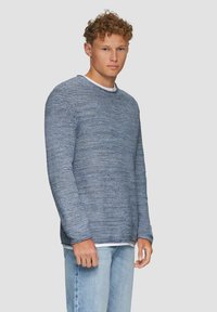 Blauwe gebreide trui met een textuurpatroon, ronde hals en lange mouwen, gelaagd over een witte shirt, gecombineerd met lichtblauwe jeans.