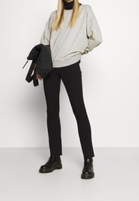 Donna che indossa leggings neri a costine, stivali neri, una felpa grigia oversize, tenendo in mano una giacca trapuntata nera piegata su uno sfondo bianco.
