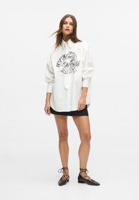 Biała, oversize'owa koszula z czarnym wzorem graficznym, wyposażona w kołnierzyk i zapinanie na guziki, zestawiona z czarną mini spódnicą i płaskimi butami ze sznurowaniem.