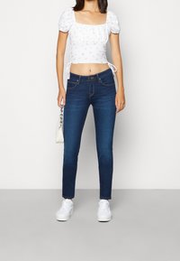 Pantalones vaqueros ajustados de mezclilla azul con cintura media, combinados con un top corto blanco con mangas abullonadas y estampado floral, y zapatillas blancas.