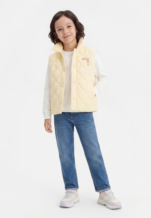 Középhosszú sötét hajú gyermek, világos sárga, steppelt mellényt, krémszínű pulóvert, kék farmert és pasztell színű sportcipőt viselve, egyik kezét a zsebében tartva áll.