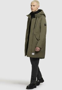 Cappotto imbottito verde oliva con cappuccio removibile, tasche laterali e zip frontale. Indossato con jeans neri e stivali neri robusti.
