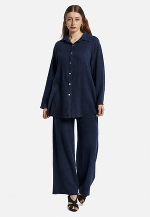 Vrouw staand, dragend een losse marineblauwe blouse met knopen en bijpassende wijde broek met zwarte open teen hakken tegen een effen lichte achtergrond.