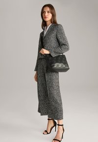 Bolso de mano negro texturizado con diseño en relieve, combinado con un traje de tweed gris y tacones negros de tiras, creando un look monocromático.