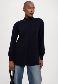 Marineblå langermet skjorte med høy krave, med knappelukking og mansjetterte ermer, kombinert med lyseblå jeans for kontrast.