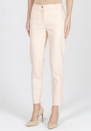 Kobieta ubrana w beżowe dopasowane eleganckie spodnie i pasujące beżowe buty na wysokim obcasie, stojąca na tle jasnego, jednolitego tła.