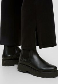 Zwarte leren enkelboots met een chunky zool, elastische zijpanelen en minimale stikdetails, gecombineerd met zwarte broeken met zijsplitten.