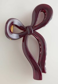 Burgundi spalvos plaukų segtukas, suformuotas kaip kaspinas, blizgiu paviršiumi, su susuktomis kilpomis ir auksine spiralės akcentu centre.