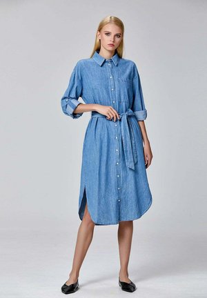 Femme portant une robe en jean bleu à boutons avec des manches retroussées, ceinture nouée à la taille, fentes sur les côtés et ballerines noires, se tenant devant un fond uni.