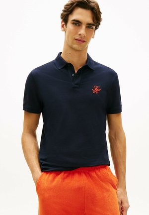 Młody mężczyzna w granatowej koszulce polo z czerwonym logo i jaskrawo pomarańczowych szortach stoi z rękami w kieszeniach na białym tle.