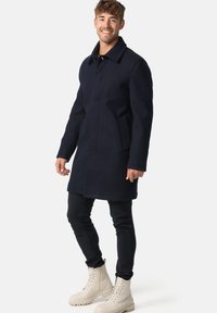 Marineblå uldblandingsfrakke med krave, forreste lommer og et enkelt design. Modellen har sorte bukser og beige snørebåndsstøvler på.