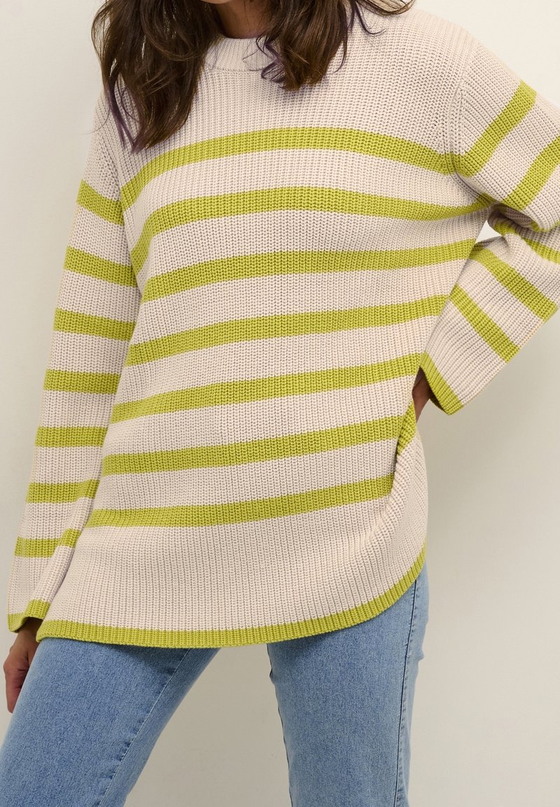 Femme portant un pull en tricot beige avec des rayures horizontales vertes et un jean bleu clair, debout avec une main sur la hanche.