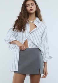 Junge Frau mit langen, lockigen Haaren, die ein weißes, gestreiftes Oversized-Hemd, einen grauen Minirock mit Schlitz vorne und eine sternförmige Halskette trägt.