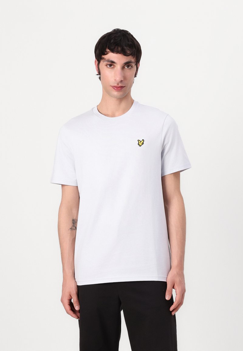 T-shirt en coton blanc à manches courtes, avec un petit logo brodé jaune sur la poitrine gauche. Porté avec un pantalon noir.