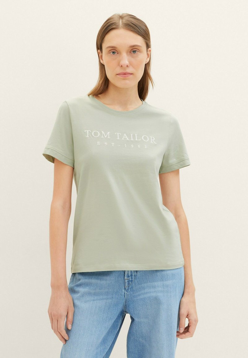 T-shirt en coton vert clair avec un col ras du cou et des manches courtes. Présente le texte "TOM TAILOR EST. 1962" en blanc sur la poitrine.