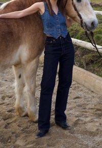 Denimweste mit Knöpfen, dunkelblaue hochgeschnittene Jeans und schwarze Stiefel. Ein hellbraunes Pferd steht daneben, vor einem sandigen Hintergrund.