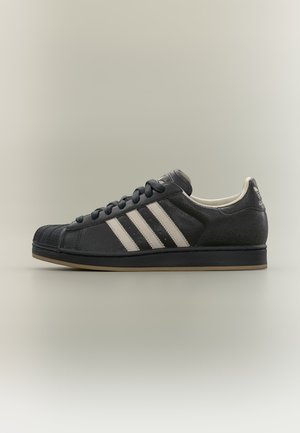 Baskets Adidas noires avec trois bandes blanches, bout en caoutchouc, lacets noirs et semelle marron sur fond neutre.