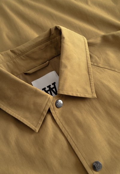 Veste beige à col avec des boutons pression argentés embossés d'un "W" et une étiquette en tissu blanc à l'intérieur du col montrant un "W" noir.