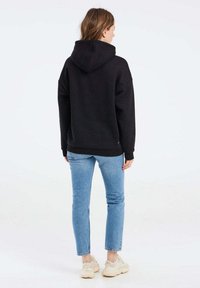 Felpa nera con cappuccio grande, vestibilità oversize, polsini a costine, indossata con jeans tapered azzurri chiari e sneakers chiare. Design minimalista.