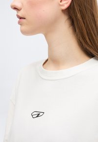 Νεαρό άτομο με ανοιχτόχρωμο δέρμα και καστανά μαλλιά, που φοράει λευκό μπλουζάκι με στρογγυλή λαιμόκοψη και μικρό μαύρο αφηρημένο λογότυπο στο στήθος.